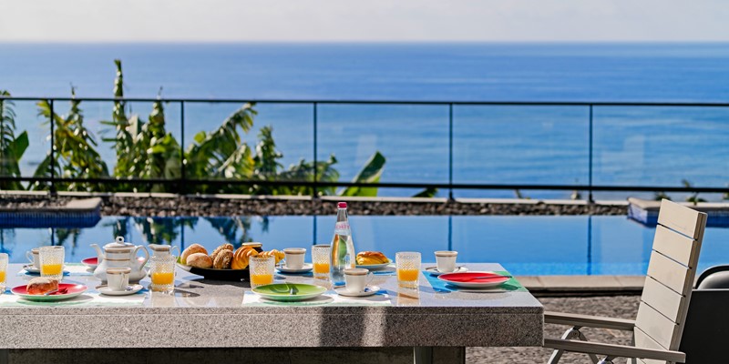 Close Up Of The Outdoor Breakfast Table At Graycis House By Ourmadeira With Fresh Food In Focus And The Swimming Pool And Ocean Softly Blurred In The Background